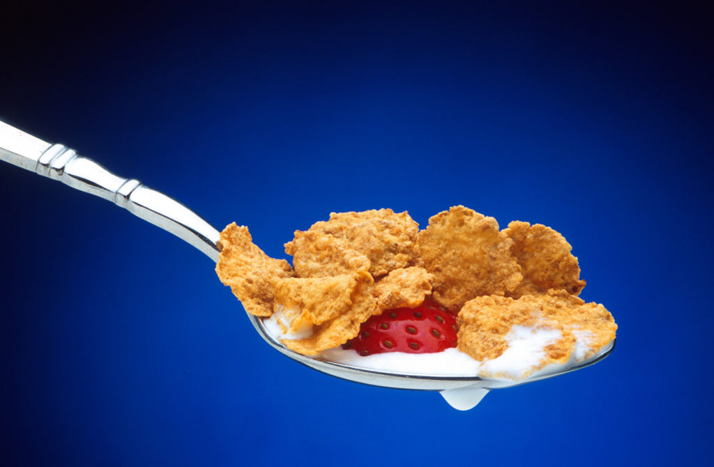 A spoon containing breakfast cereal flakes, part of a strawberry, and milk is held in midair against a blue background