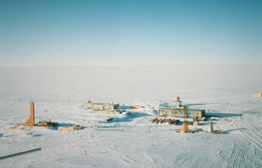 A photo of Wostok station in Antartica. Set against an otherwise barren snowy landscape, several small buildings resembling shipping containers are grouped together, with smaller constructions around them.