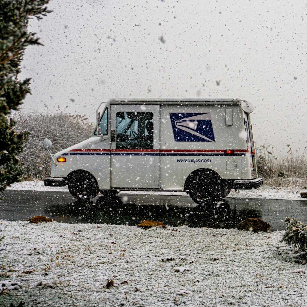 A photo of a USPS truck driving on the road as snow falls