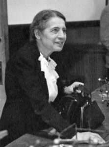 A photograph of Lise Meitner, after whom meitnerium was named. She leans forward against a desk as she intensely engages someone off off-camera.