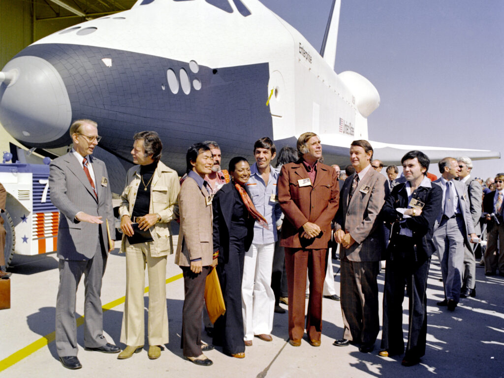 The Shuttle Enterprise rolls out of the Palmdale manufacturing facilities with Star Trek television cast members. From left to right they are: Dr. James C. Fletcher (NASA Administrator), DeForest Kelley (Dr. "Bones" McCoy), George Takei (Mr. Sulu), James Doohan (Chief Engineer Montgomery "Scotty" Scott), Nichelle Nichols (Lt. Uhura), Leonard Nimoy (the indefatigable Mr. Spock), Gene Roddenberry (The Great Bird of the Galaxy), Democratic Congressman Don Fuqua, and Walter Koenig (Ensign Pavel Chekov).