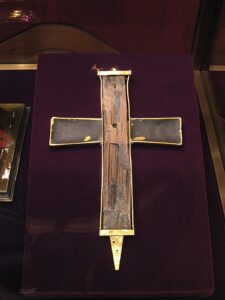 A photo of a fragment of the true cross, held for veneration in Vienna. The wood, is held in a metal and glass case in the shape of cross, laid against a red velvet cushion